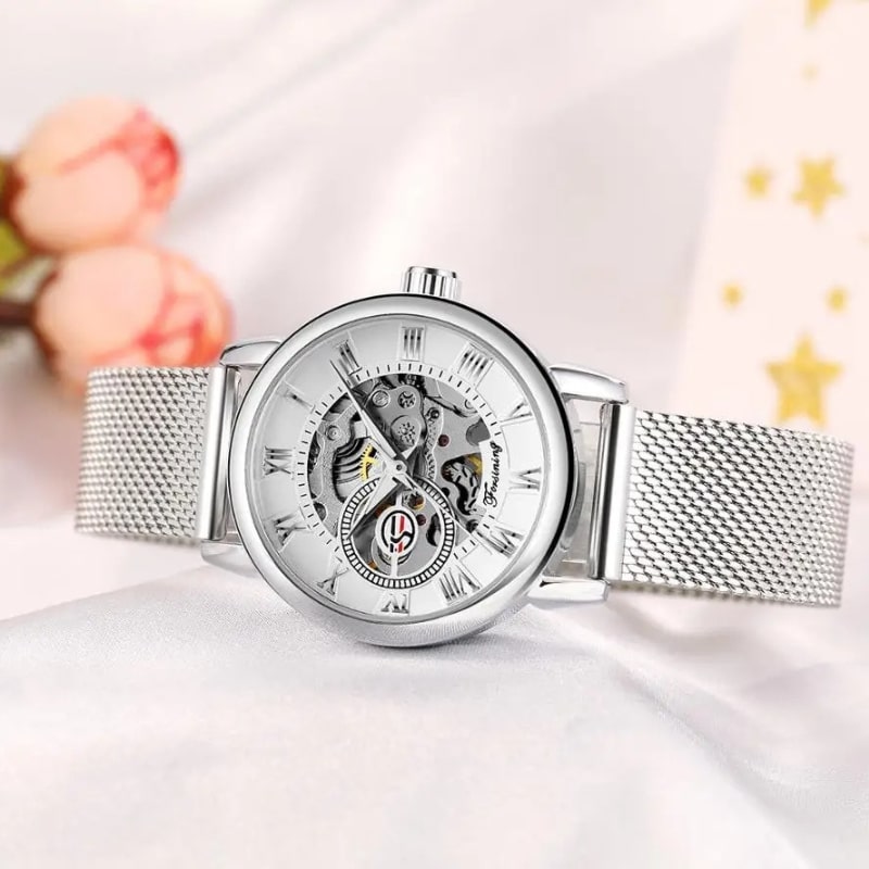 Montre Automatique Femme Bracelet Acier Maille & Cadran Ajouré en argenté couchée sur une table avec des fleurs en arrière plan